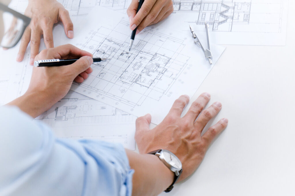 Zwei Personen arbeiten gemeinsam an einem Architekturplan mit Fokus auf die Lichtplanung in einem Bürogebäude. Auf dem Tisch liegen technische Zeichnungen mit markierten Leuchtpunkten, ein Zirkel und Schreibutensilien – typische Werkzeuge für präzise Lichtkonzepte im Innenraum.