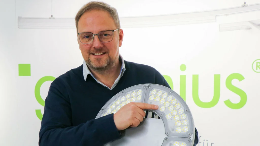 Geschäftsführer der greenius GmbH & Co. KG präsentiert ein LED-Modul mit zahlreichen Einzel-LEDs und zeigt auf die Technik. Im Hintergrund ist das Firmenlogo „greenius“ sowie ein Lichtbandsystem zu sehen, was den Fokus auf innovative LED-Lösungen unterstreicht.